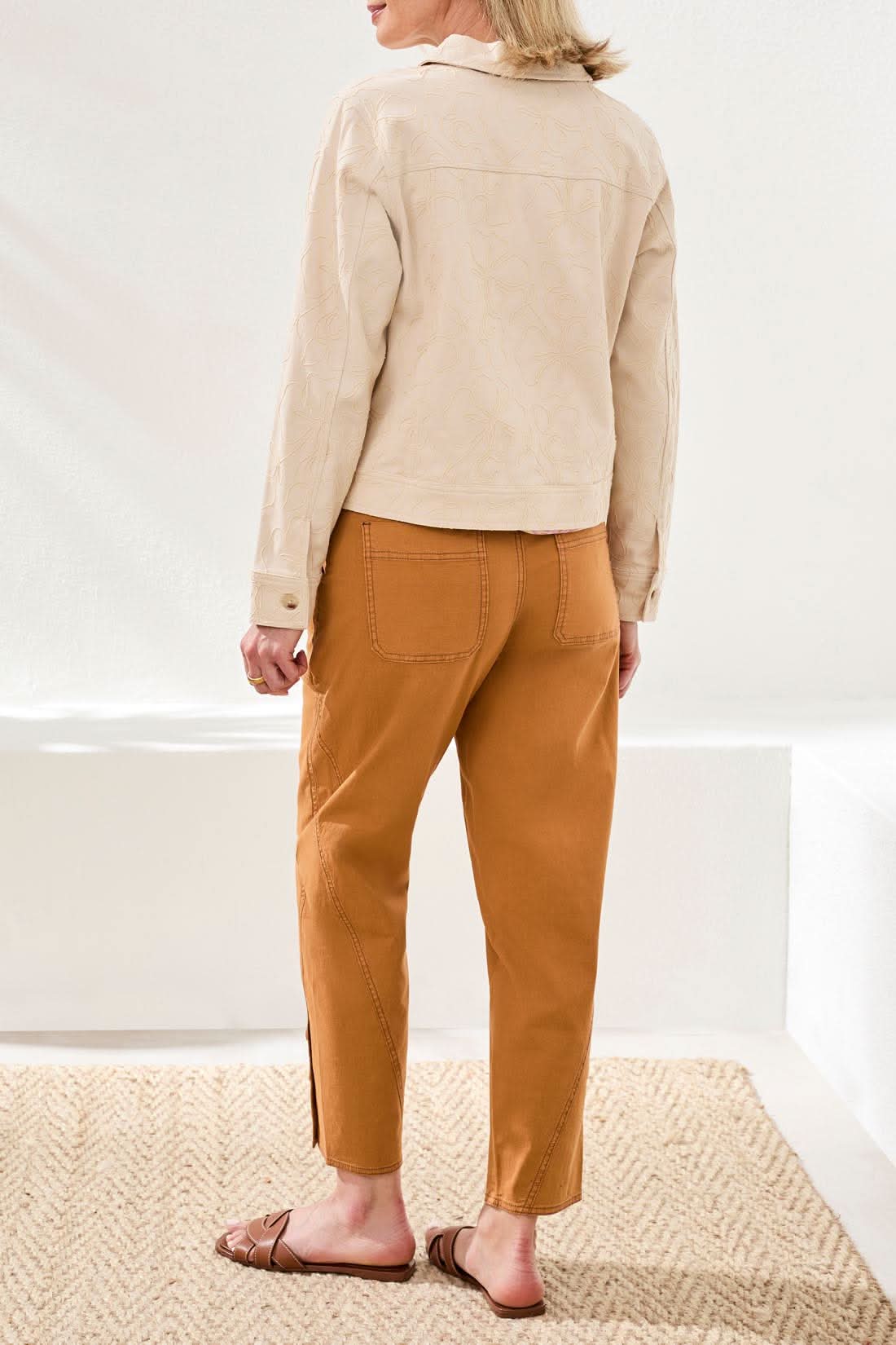 A woman wears the Tribal Floral Textured Jacket, a light beige, long-sleeve button-front piece with front pockets and floral embroidery, styled with high-waisted brown pants.