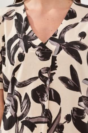 A woman stands indoors wearing the Tribal TUNIC DRESS, a breathable cotton, knee-length cream summer dress with black leafy print and short sleeves, paired with black flip-flops.