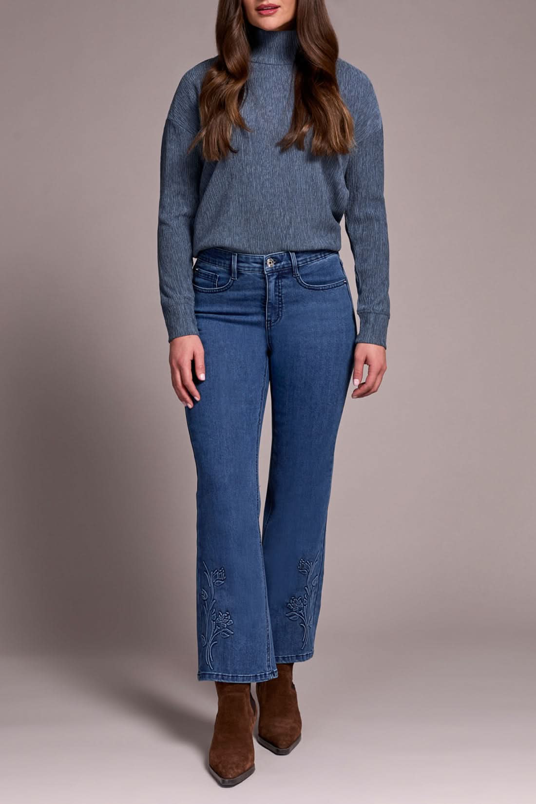 A woman in a gray ribbed turtleneck sweater, Tribal AUDREY FLARE JEANS featuring a floral hem design, and brown suede ankle boots.