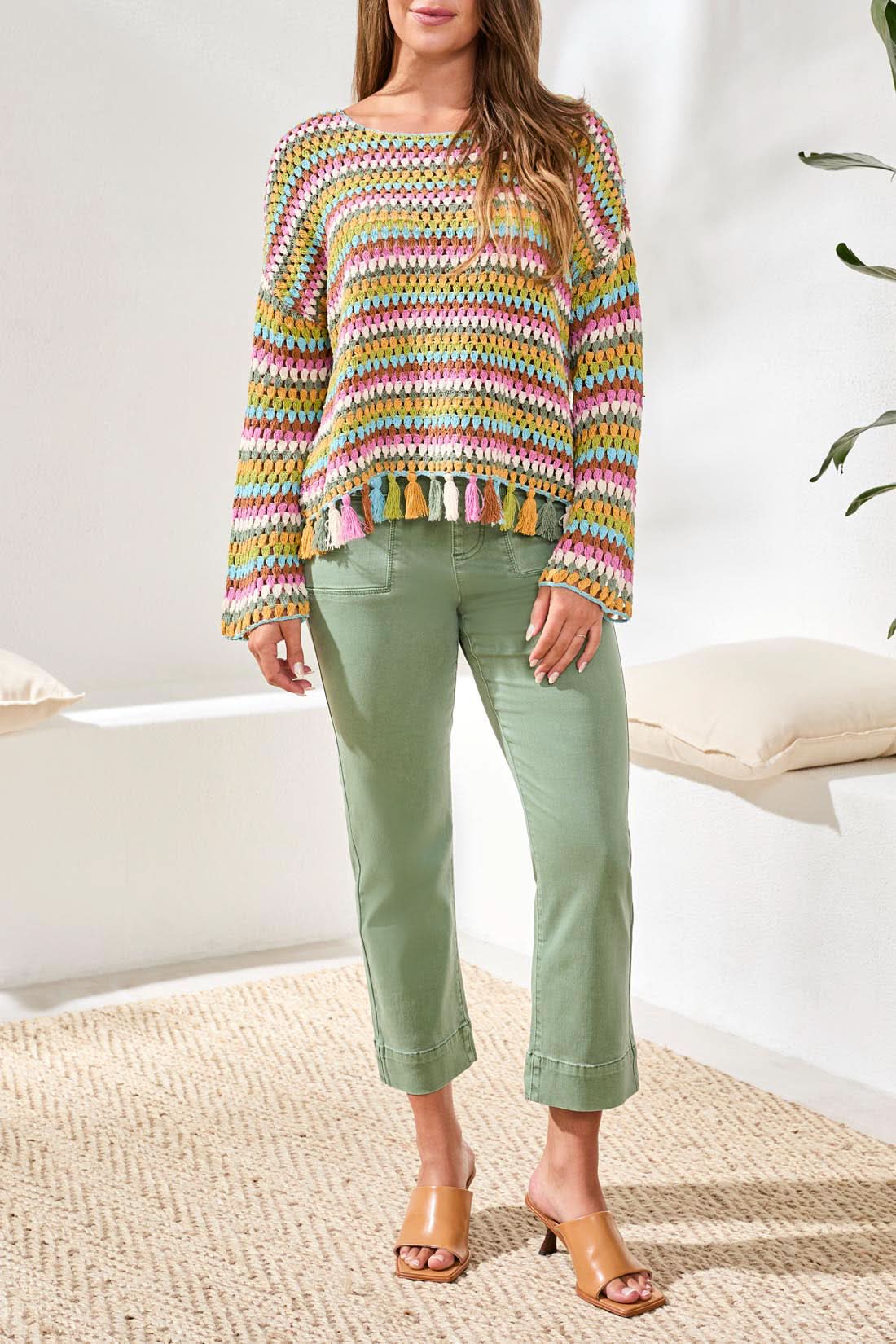 A woman smiles at the camera in front of a light blue door, wearing the Tribal Crochet Bell Sleeve Top with tassels and pink pants.
