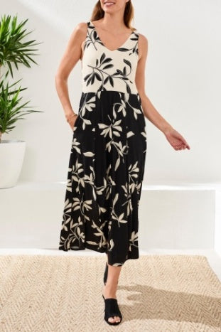 A woman stands indoors on a woven rug, wearing the Tribal SLVLESS COMBO DRESS with a V-neck and black-and-white floral print, featuring a flowy silhouette. She pairs it with black open-toe shoes.