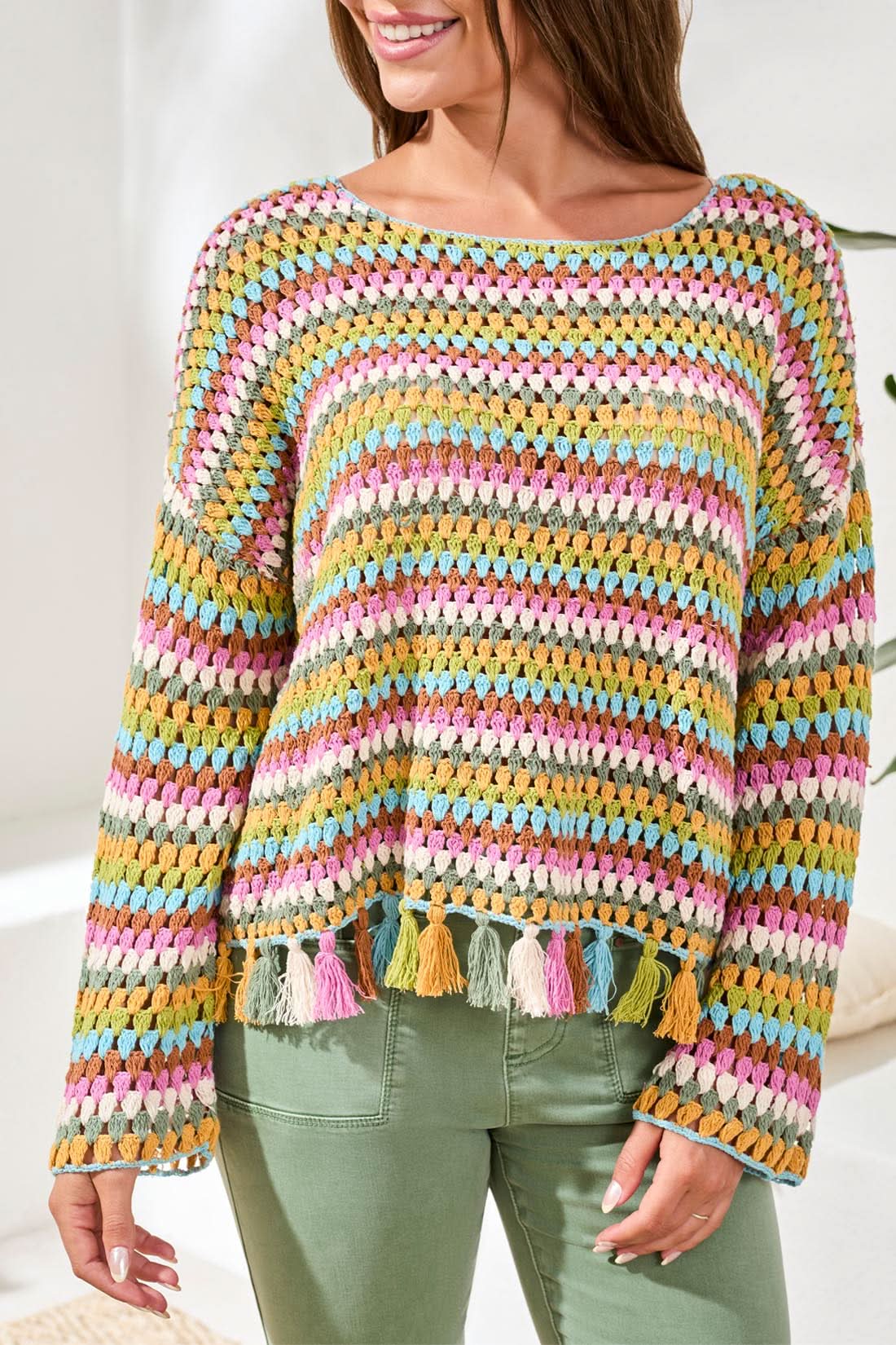 A woman smiles at the camera in front of a light blue door, wearing the Tribal Crochet Bell Sleeve Top with tassels and pink pants.