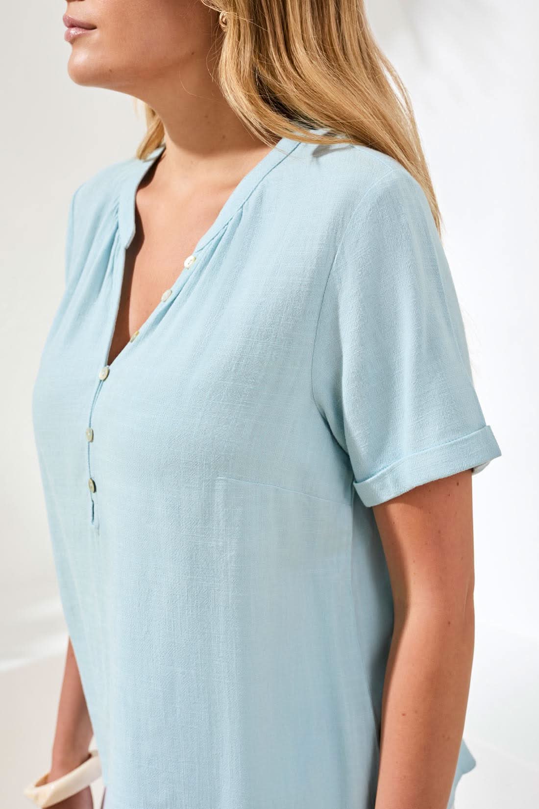 A woman wears the Tribal Chic Coastal Shirt—a light blue, short sleeve linen-rayon blouse with buttons—shown from shoulders to mid-torso against a white background.