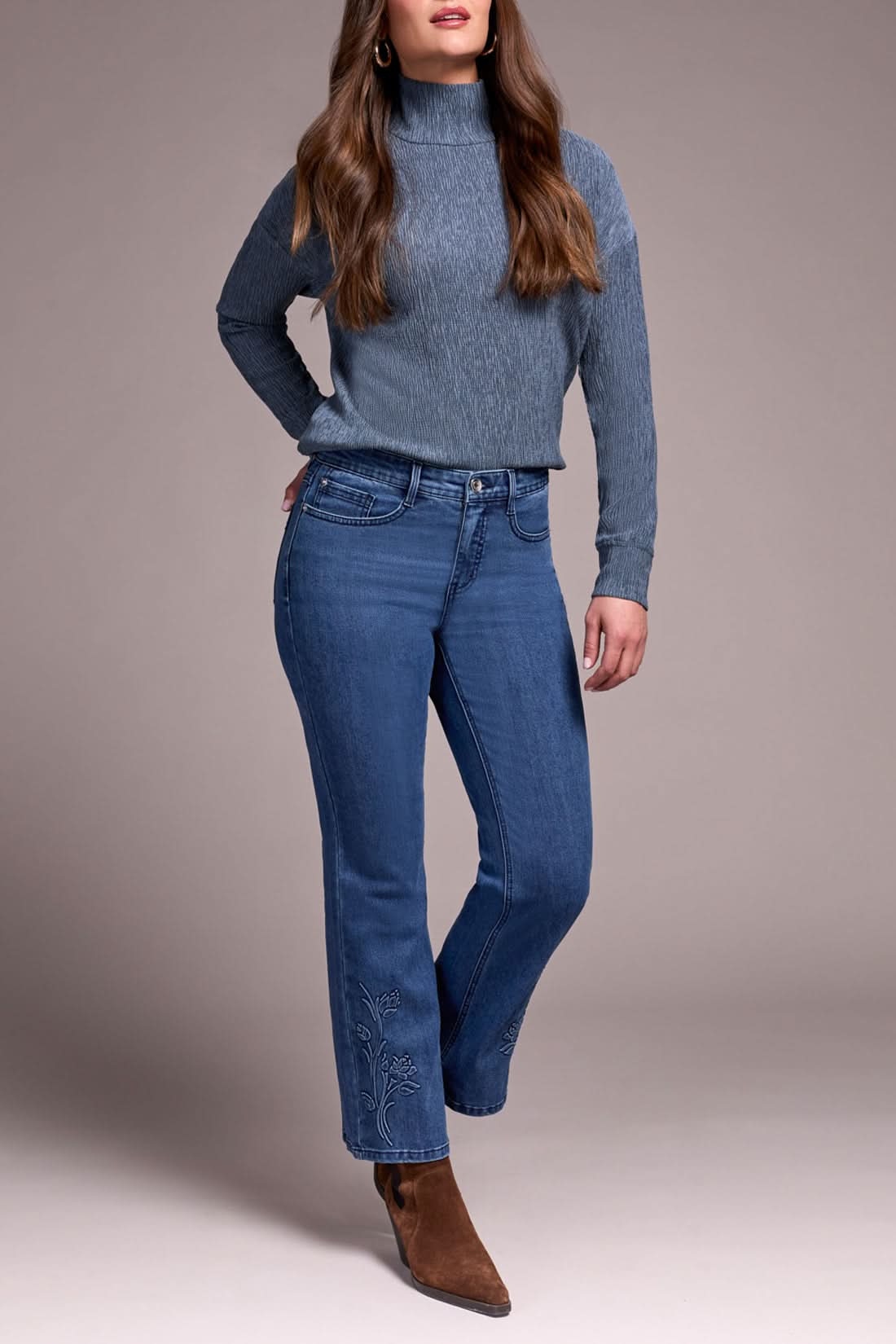 A woman in a gray ribbed turtleneck sweater, Tribal AUDREY FLARE JEANS featuring a floral hem design, and brown suede ankle boots.