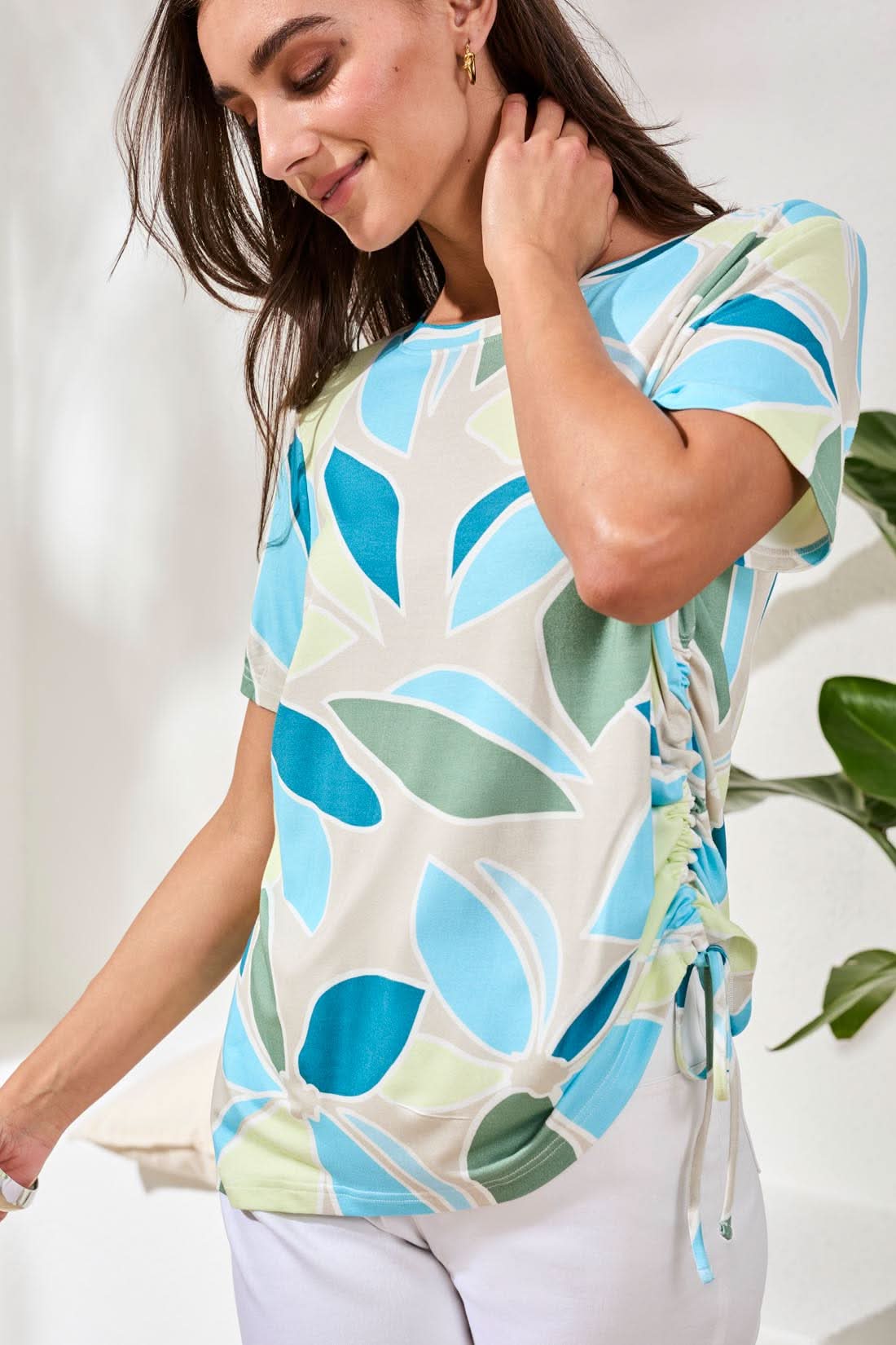A woman wearing a Tribal Boat Neck Top in aqua sky with side ruching and white pants stands indoors near a cushion.