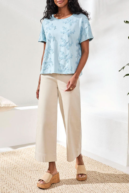 A person sits on a white surface wearing the Tribal Design Crew Neck Top in light blue with a floral textured pattern and light beige pants, showcasing nature-inspired fashion.