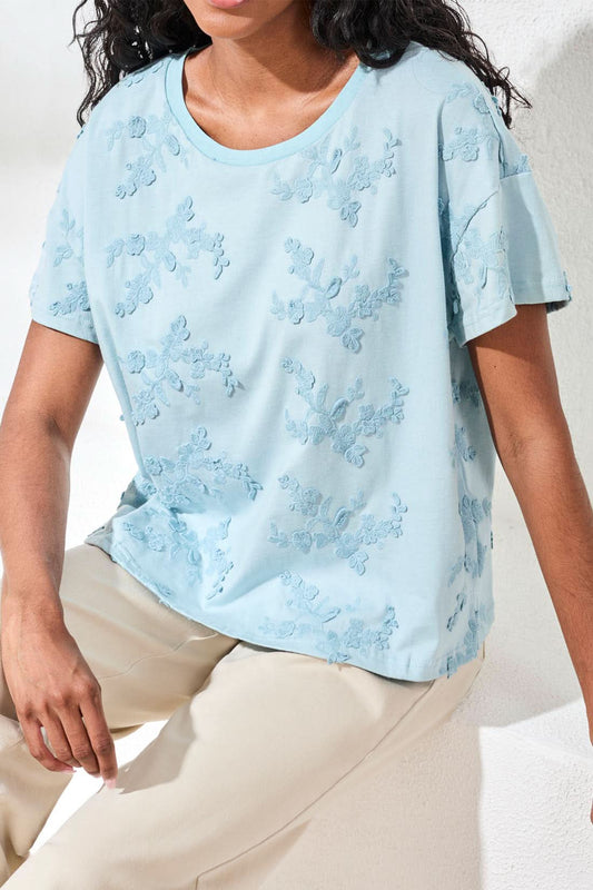 A person sits on a white surface wearing the Tribal Design Crew Neck Top in light blue with a floral textured pattern and light beige pants, showcasing nature-inspired fashion.