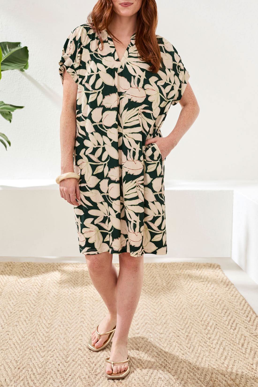 A woman models the Tribal Ruffle Mid-length Dress featuring a relaxed shift silhouette and rainforest-inspired beige and dark green leaf print, standing on a woven rug with one hand in her pocket.
