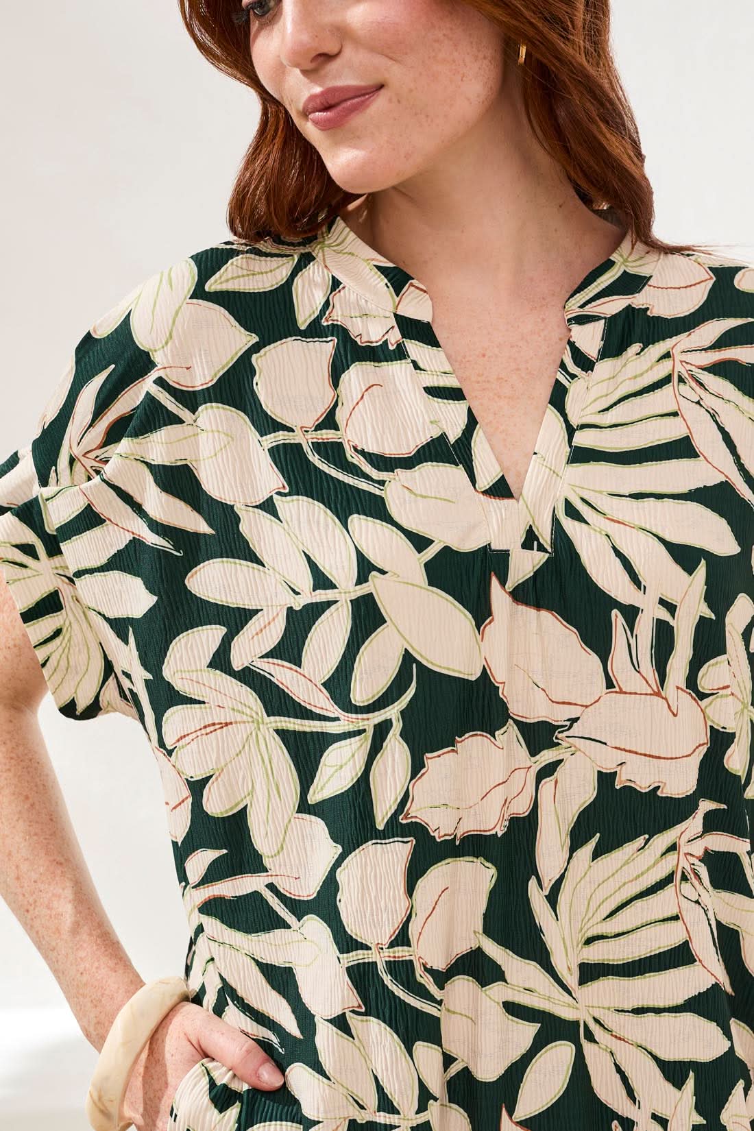 A woman models the Tribal Ruffle Mid-length Dress featuring a relaxed shift silhouette and rainforest-inspired beige and dark green leaf print, standing on a woven rug with one hand in her pocket.
