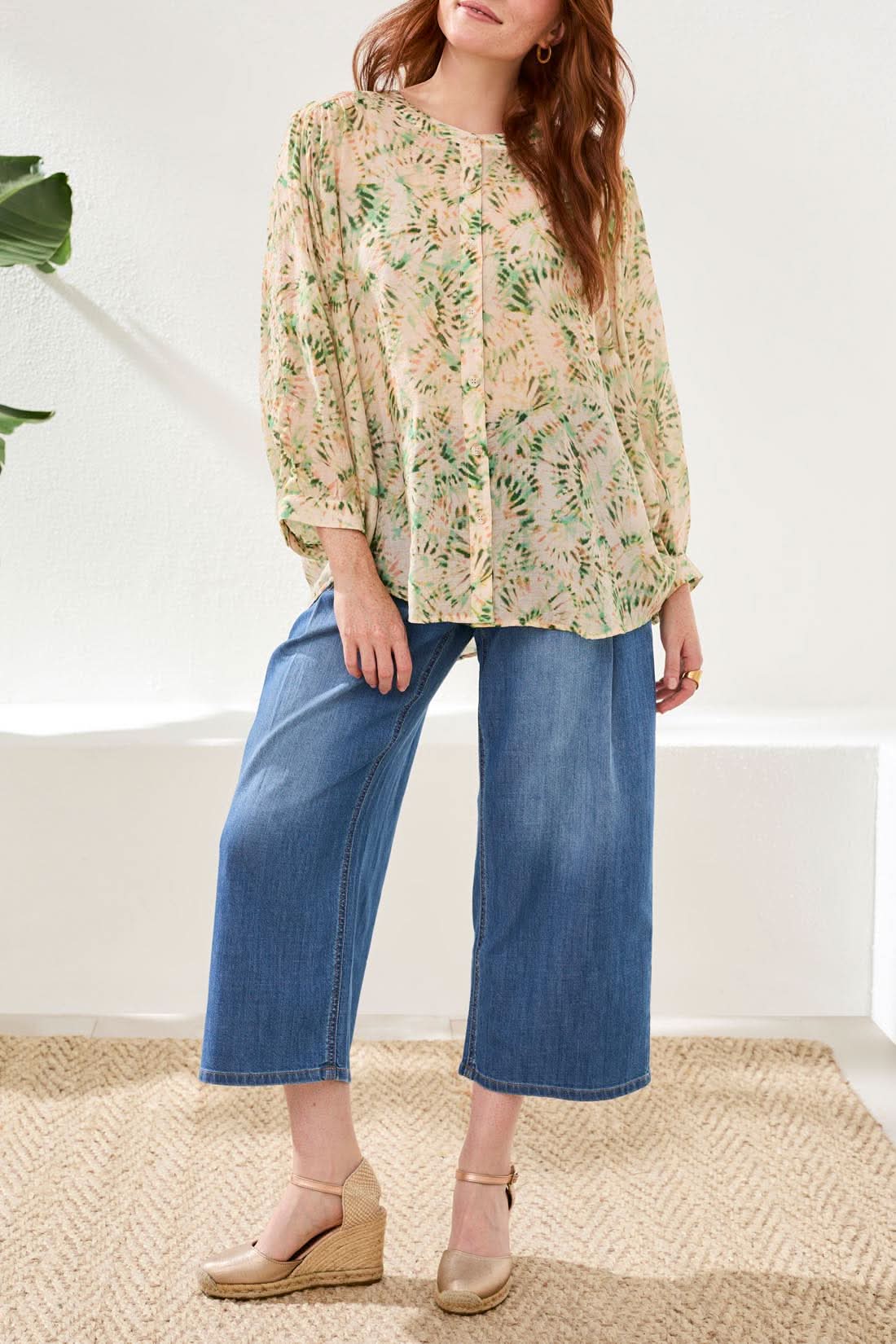 A woman wears the Tribal Dolman Sleeve Blouse, paired with wide-leg blue jeans and beige wedge sandals, standing on a woven rug against a white background.