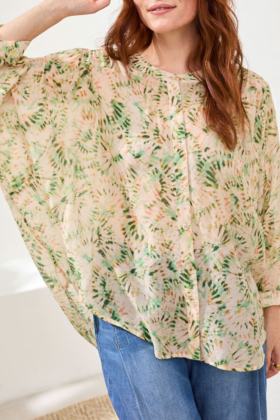 A woman wears the Tribal Dolman Sleeve Blouse, paired with wide-leg blue jeans and beige wedge sandals, standing on a woven rug against a white background.
