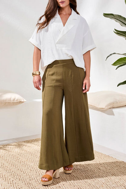 Indoors, a woman wears Tribal Wide Leg Flowy Pant in olive green, paired with a loose white short-sleeve blouse, tan sandals, and a gold bracelet.