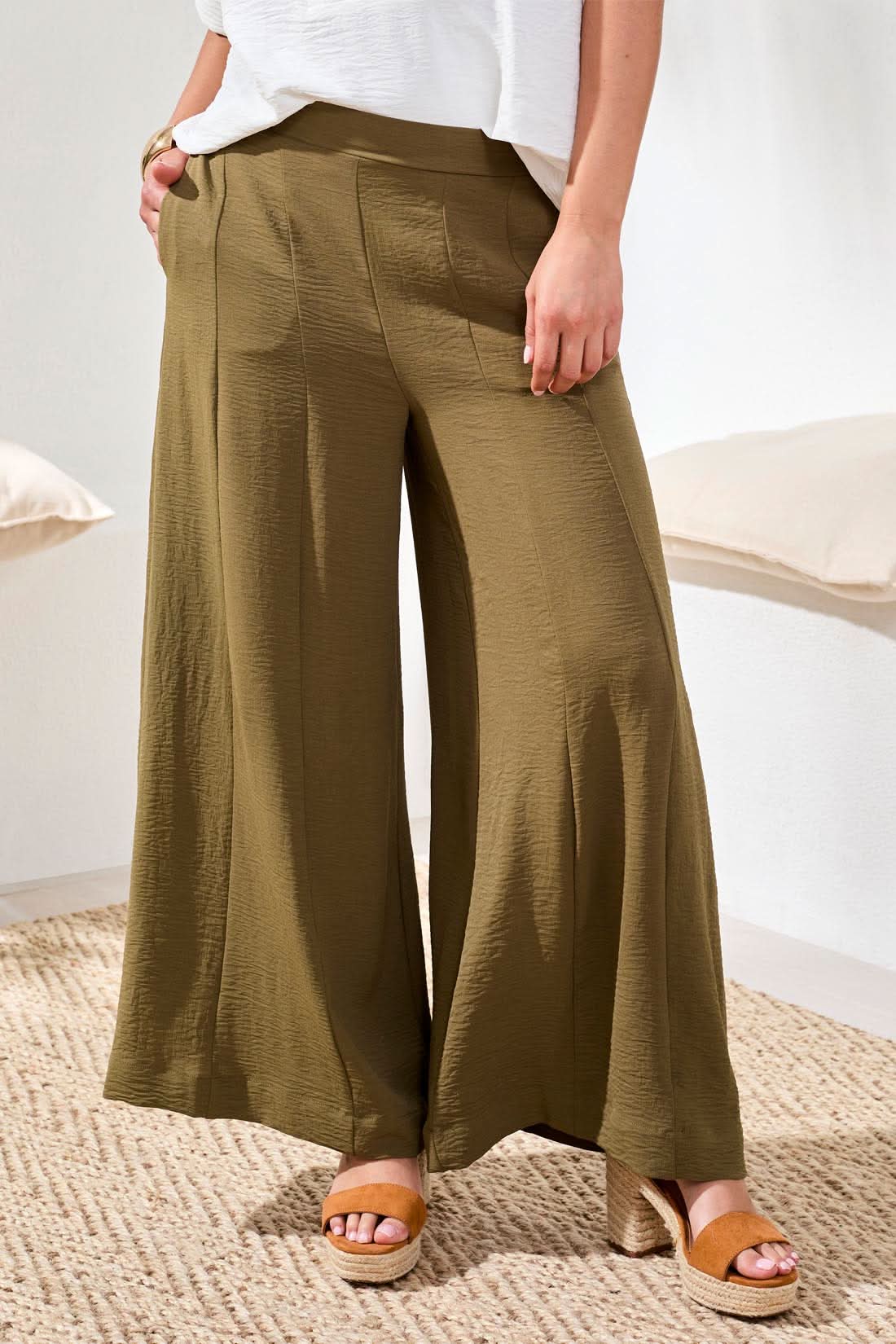 Indoors, a woman wears Tribal Wide Leg Flowy Pant in olive green, paired with a loose white short-sleeve blouse, tan sandals, and a gold bracelet.