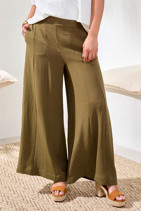 Indoors, a woman wears Tribal Wide Leg Flowy Pant in olive green, paired with a loose white short-sleeve blouse, tan sandals, and a gold bracelet.