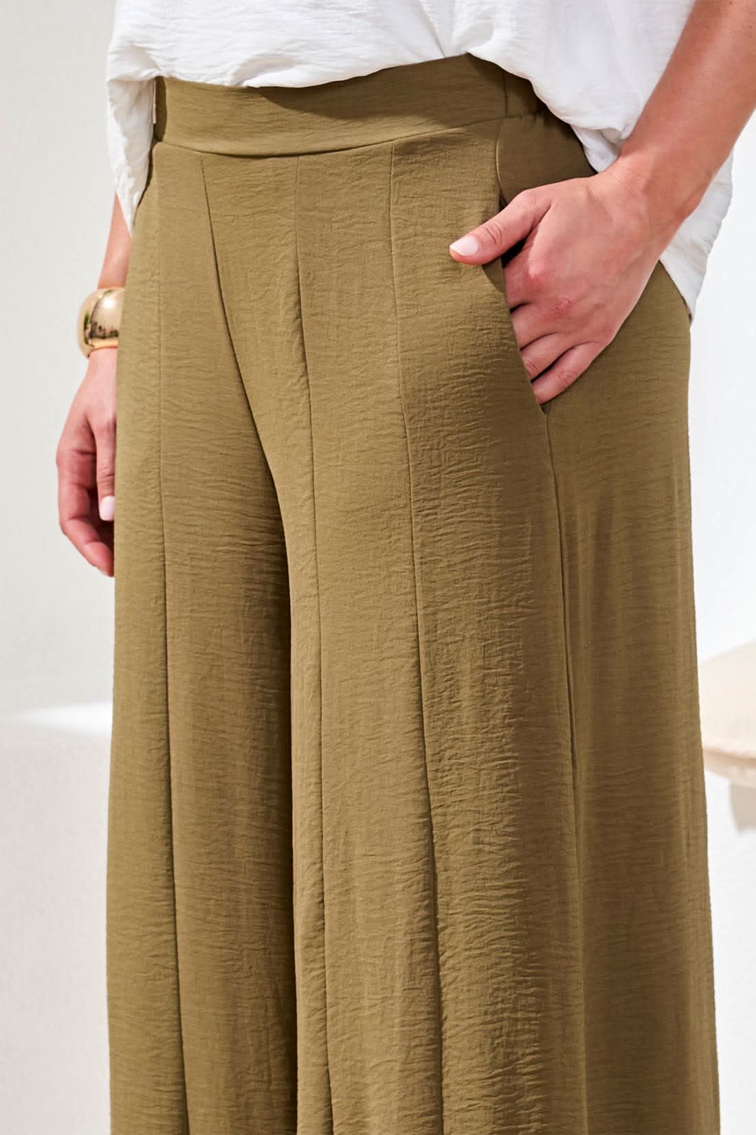 Indoors, a woman wears Tribal Wide Leg Flowy Pant in olive green, paired with a loose white short-sleeve blouse, tan sandals, and a gold bracelet.
