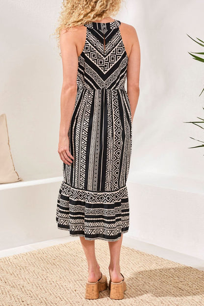 A woman stands against a stone wall, wearing the Tribal HALTER DRESS—sleeveless with a black and white pattern—and black sandals, her hair down, one hand in her pocket.