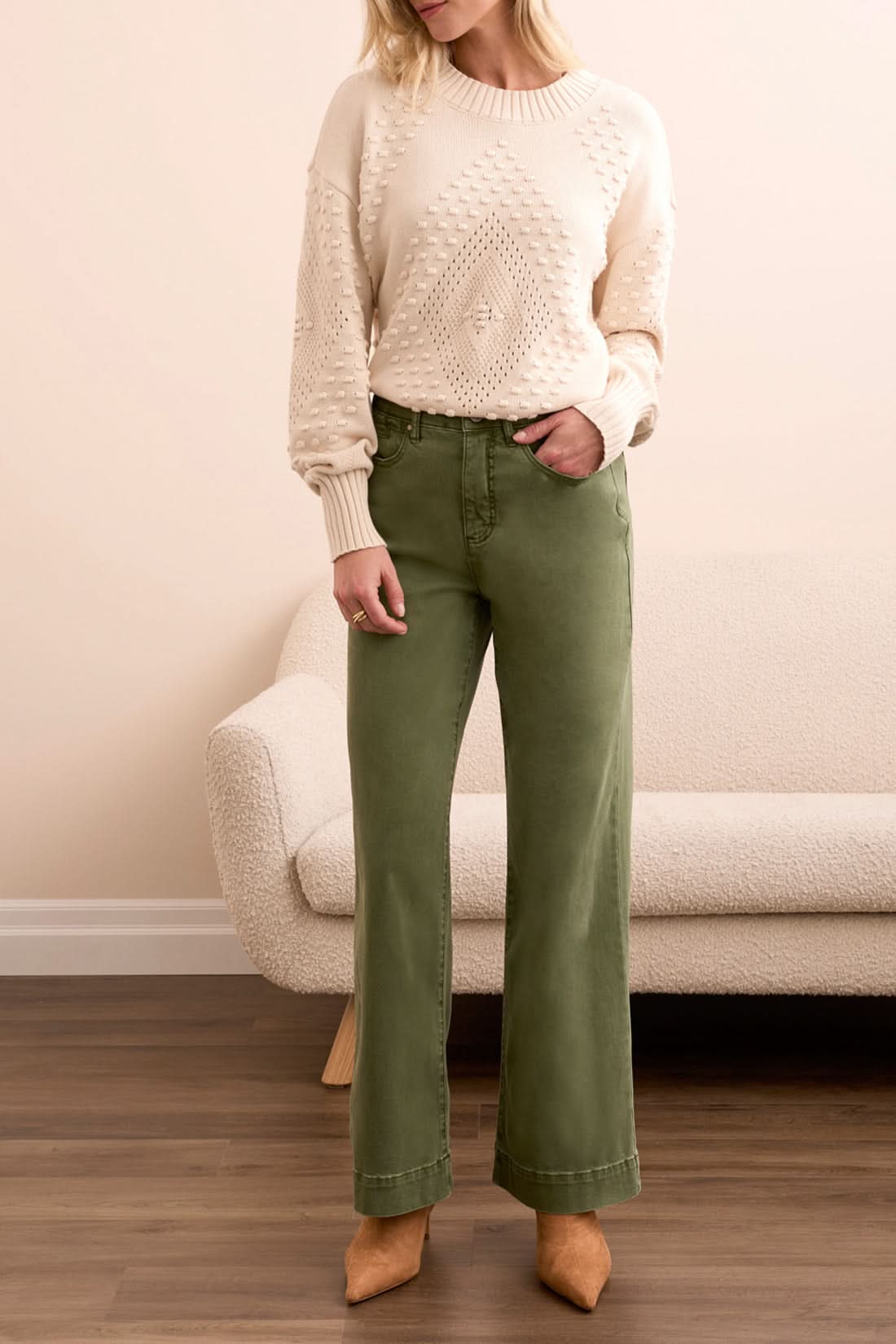 A woman stands indoors in Tribal FLY FRONT WIDE LEG PANTS, a cream knit sweater, and tan heels, posed by a light sofa on a wooden floor.