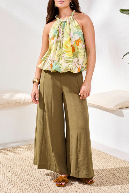 A woman stands indoors on a woven rug, wearing Tribal's Bubble Blouse—a sleeveless halter top in 100% cotton with a floral print—paired with olive wide-leg pants.