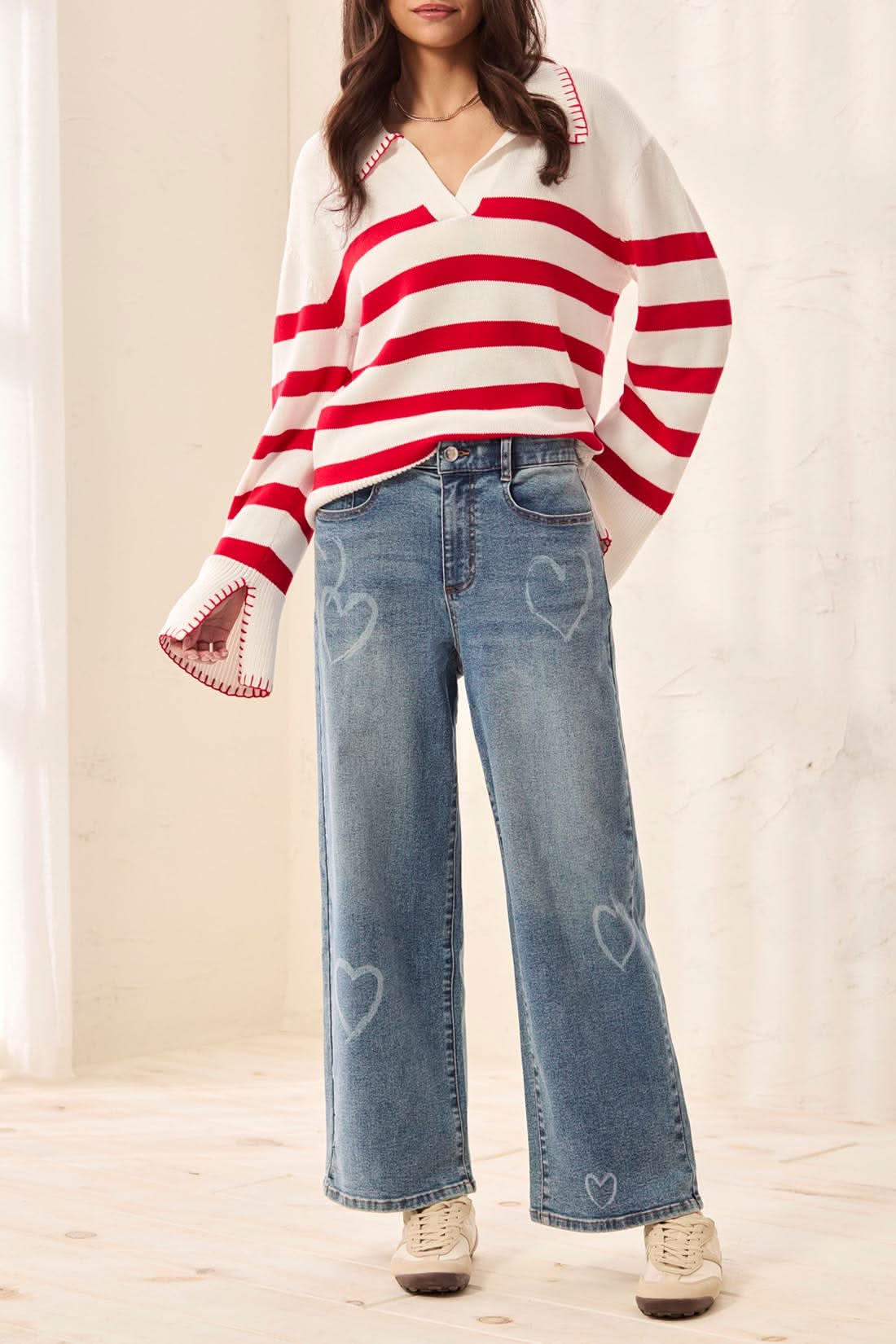 A person indoors wearing Tribal’s Sophia Printed Straight Jeans with faint heart patterns, a red and white striped sweater, and white sneakers.