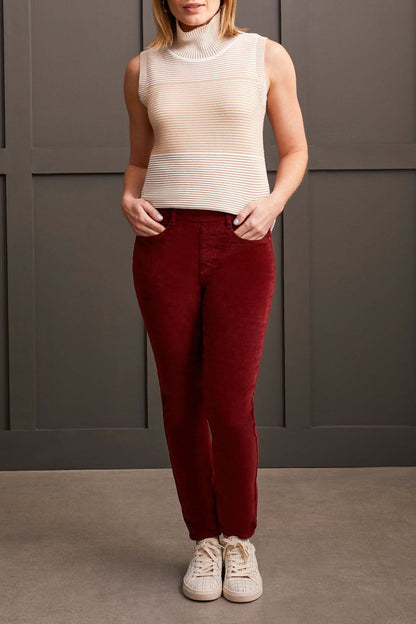 A person clad in a cream-colored quilted coat, a striped top, Tribal Audrey Pull on Slim Ankle Pants in burgundy, and white sneakers stands against a paneled grey wall.