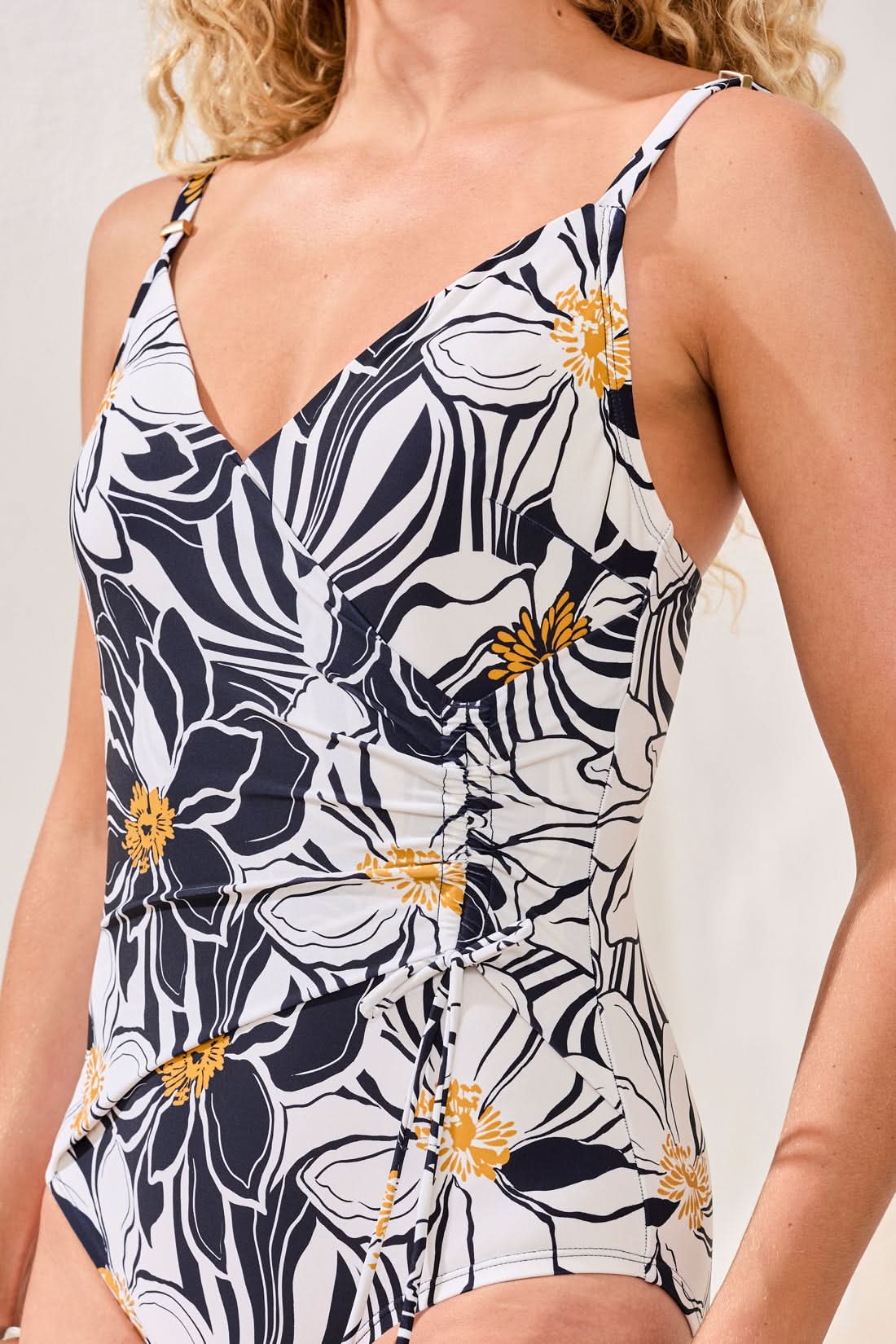 A woman models the Tribal Wrap Front Swimsuit, featuring a black and white floral print with yellow accents. She stands indoors on a woven rug, beige cushions and a green plant in the background.