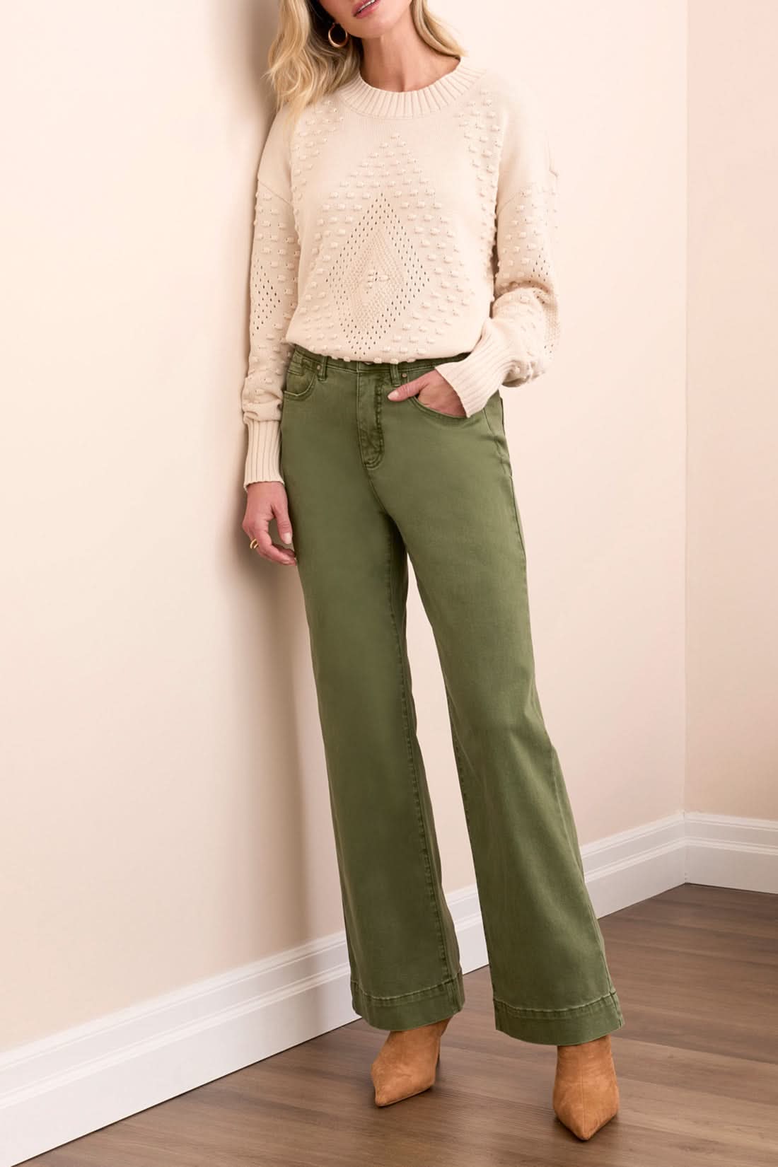 A woman stands indoors in Tribal FLY FRONT WIDE LEG PANTS, a cream knit sweater, and tan heels, posed by a light sofa on a wooden floor.