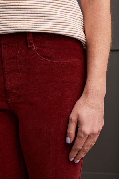 A person clad in a cream-colored quilted coat, a striped top, Tribal Audrey Pull on Slim Ankle Pants in burgundy, and white sneakers stands against a paneled grey wall.