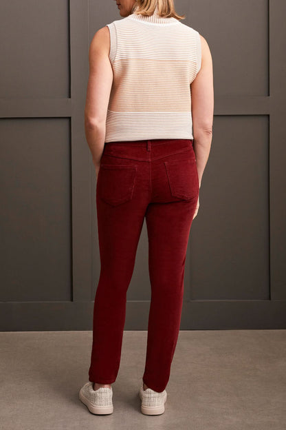 A person clad in a cream-colored quilted coat, a striped top, Tribal Audrey Pull on Slim Ankle Pants in burgundy, and white sneakers stands against a paneled grey wall.