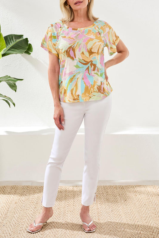 A woman wearing the Tribal Scoop Neck Flare shirt in a pastel floral pattern and white pants sits on a white surface, smiling with her head slightly turned.