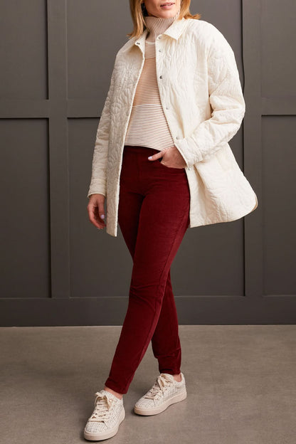 A person clad in a cream-colored quilted coat, a striped top, Tribal Audrey Pull on Slim Ankle Pants in burgundy, and white sneakers stands against a paneled grey wall.