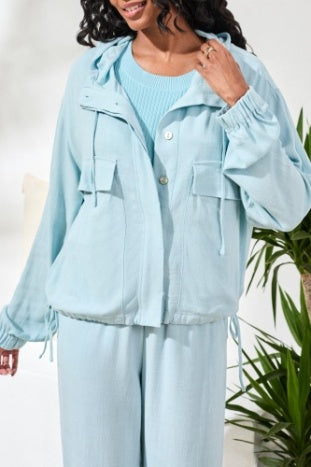 A woman wears the Tribal Light Jacket with Hood, a light blue button-up style paired with matching pants, ideal for effortless layering near green plants in a bright indoor space.