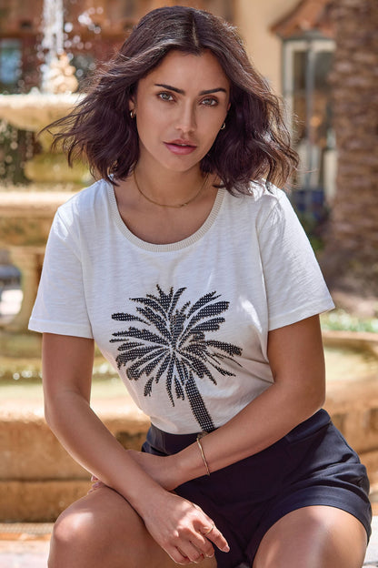 A woman with wavy dark hair sits outdoors by a fountain, wearing the Tribal CREW NECK TOP WITH PALM PRINT and black shorts.