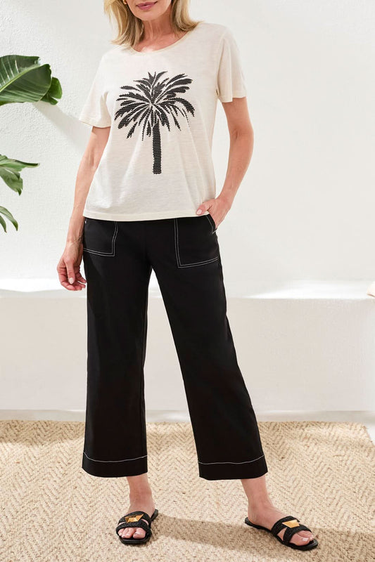 A woman with wavy dark hair sits outdoors by a fountain, wearing the Tribal CREW NECK TOP WITH PALM PRINT and black shorts.