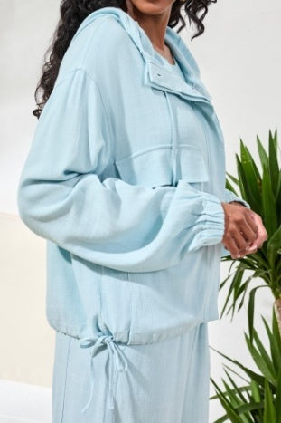 A woman wears the Tribal Light Jacket with Hood, a light blue button-up style paired with matching pants, ideal for effortless layering near green plants in a bright indoor space.