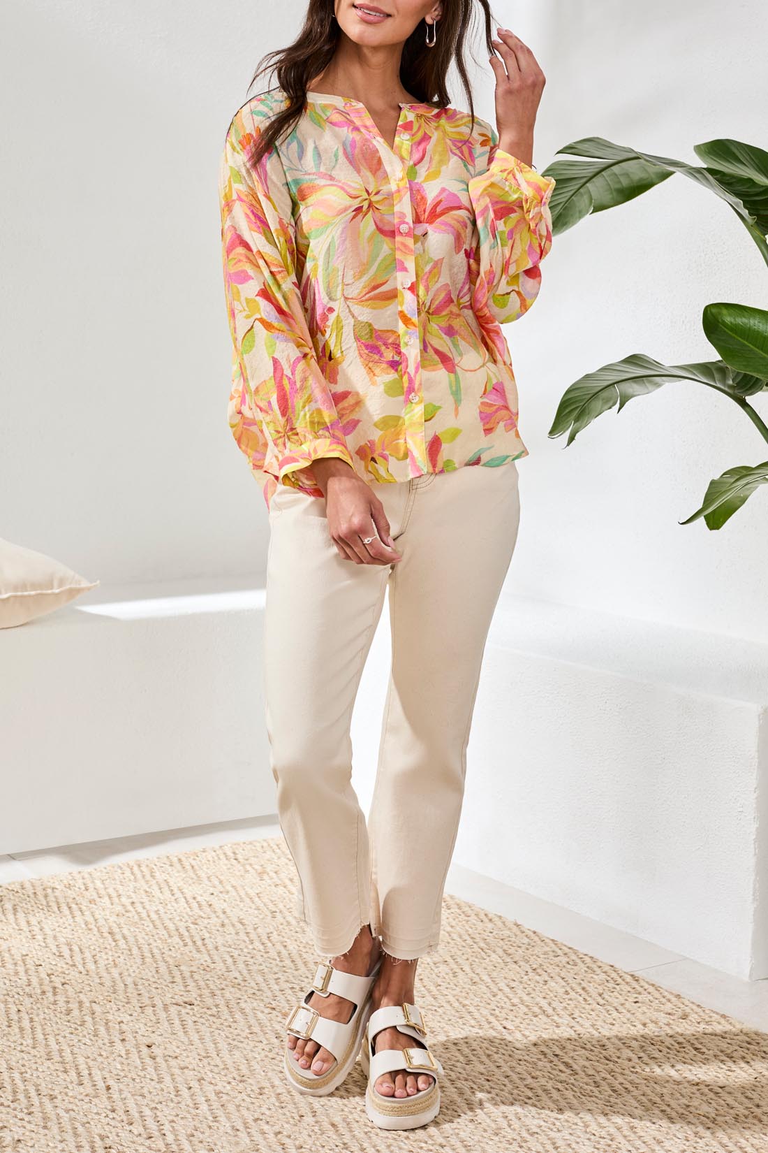 A woman with long blonde hair wears the Tribal Breezy Summer Blouse while sitting outdoors and smiling. Colorful cushions and greenery can be seen in the background.