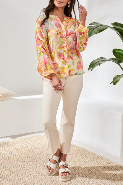 A woman with long blonde hair wears the Tribal Breezy Summer Blouse while sitting outdoors and smiling. Colorful cushions and greenery can be seen in the background.