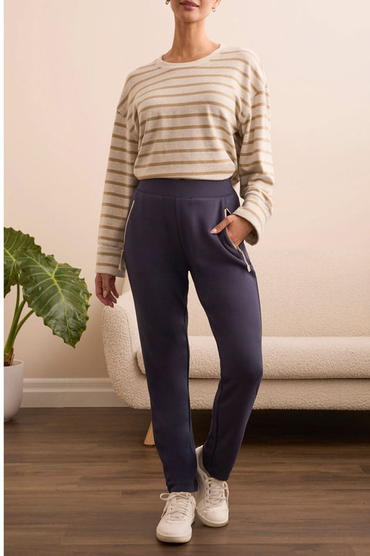 Indoors, a person stands near a potted plant and cream sofa wearing a beige striped long-sleeve shirt, white sneakers, and Tribal CONTRAST TRIM PULL-ON ANKLE PANTS with one hand in their pocket.