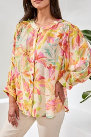 A woman with long blonde hair wears the Tribal Breezy Summer Blouse while sitting outdoors and smiling. Colorful cushions and greenery can be seen in the background.