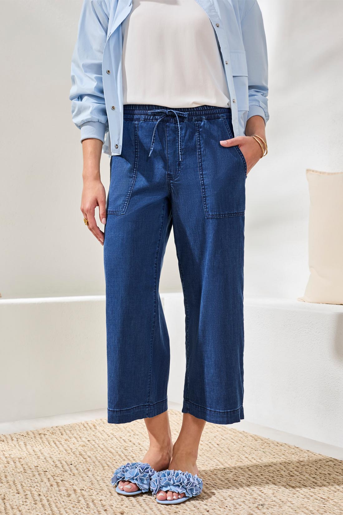 A woman stands indoors on a woven rug, wearing the Sophia Anke Jean by Tribal paired with a light blue jacket, white top, and ruffle-detailed sandals.