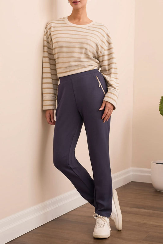 Indoors, a person stands near a potted plant and cream sofa wearing a beige striped long-sleeve shirt, white sneakers, and Tribal CONTRAST TRIM PULL-ON ANKLE PANTS with one hand in their pocket.
