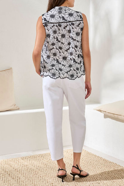 A woman stands indoors, facing away, wearing the Tribal Sleeveless Button Front Blouse with black and white floral prints, paired with white pants and black sandals. She is on a woven rug next to white benches with beige cushions.