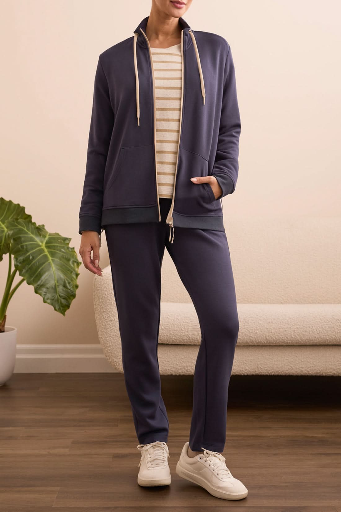 A person stands indoors in a navy tracksuit paired with the Tribal MOCK NECK CARDIGAN in beige and white stripes and white sneakers, with a green plant and cream-colored sofa in the background.