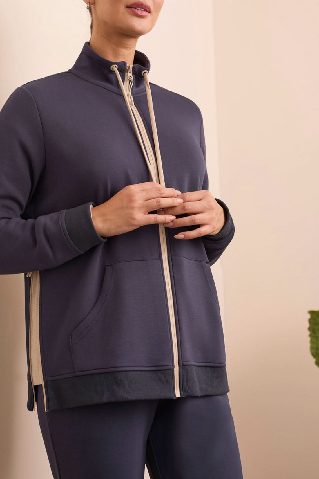 A person stands indoors in a navy tracksuit paired with the Tribal MOCK NECK CARDIGAN in beige and white stripes and white sneakers, with a green plant and cream-colored sofa in the background.