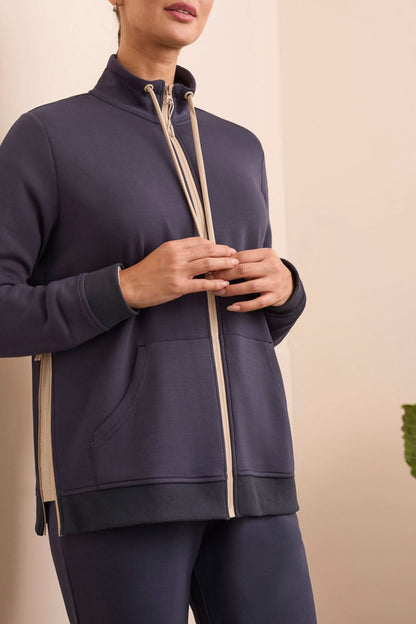 A person stands indoors in a navy tracksuit paired with the Tribal MOCK NECK CARDIGAN in beige and white stripes and white sneakers, with a green plant and cream-colored sofa in the background.