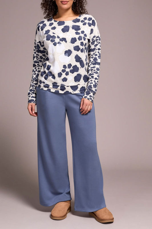 Someone stands against a neutral background wearing the Tribal CREW NECK TOP with a white and navy animal print, wide-leg blue pants, and tan slippers—an ideal look for casual wear.