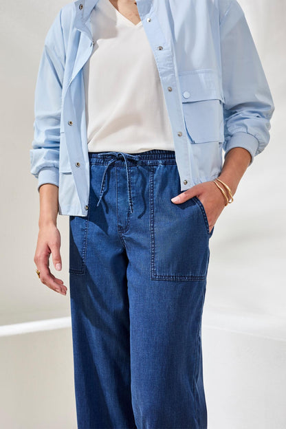 A woman stands indoors on a woven rug, wearing the Sophia Anke Jean by Tribal paired with a light blue jacket, white top, and ruffle-detailed sandals.