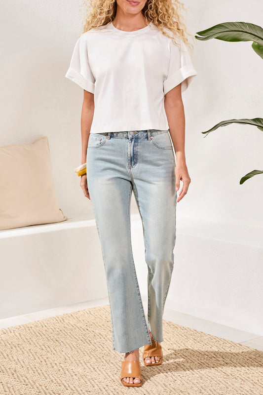 A woman stands indoors on a woven rug near a white bench and green plant, wearing the Tribal Mercerized Cotton Crew Neck T-Shirt in white, light blue straight-leg jeans, and tan heeled sandals.