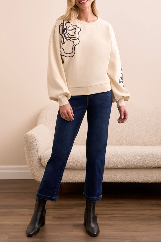 Indoors, a woman wears Tribal’s CREW NECK TOP W/EMBROIDERY—cream with black floral details and balloon sleeves—paired with dark blue jeans and black ankle boots beside a beige textured sofa.