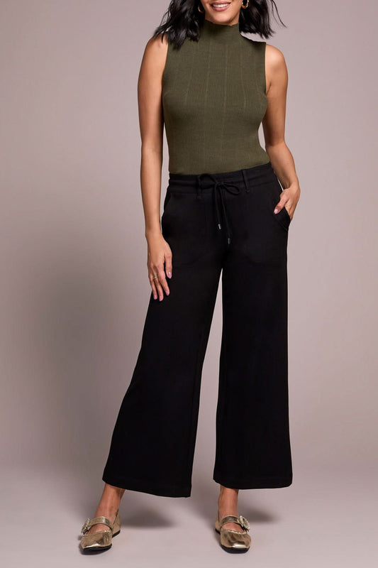 A person wearing a sleeveless olive green mock neck top, Tribal Dark Ivy Drawcord Pant in black, and gold loafers stands against a plain background.