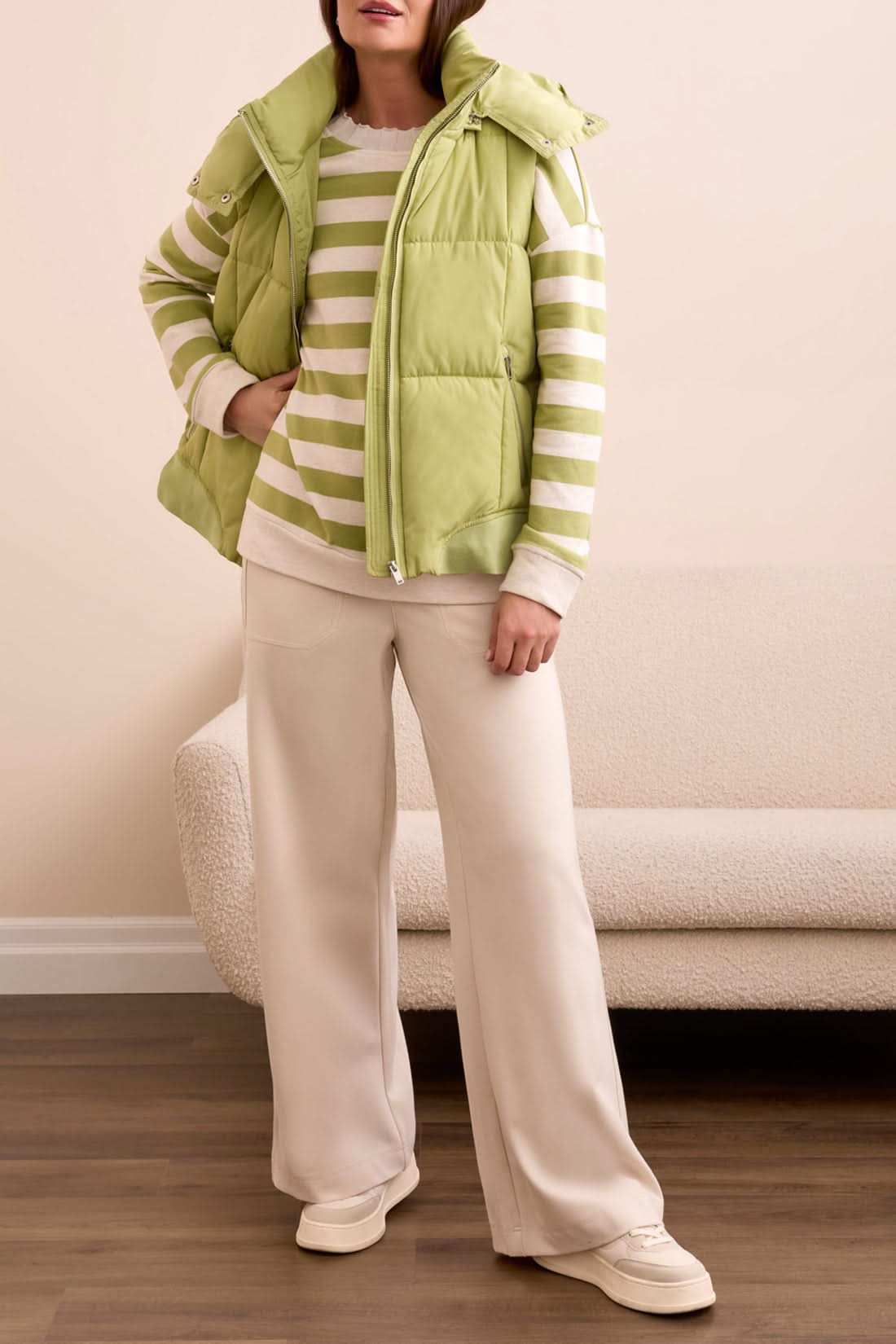 A woman stands with her hands in her pockets, looking down, wearing the Tribal QUILTED VEST in wasabi over a green and white striped sweatshirt, set against a patterned tile background.